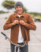 Man wearing a brown jacket and gray beanie standing next to a bicycle outdoors.