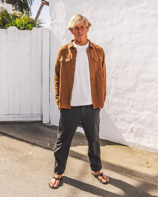 Man wearing a brown jacket, white shirt, and dark pants standing against a white wall.