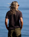 Person with long hair and sunglasses standing by a body of water wearing a dark shirt and green pants.