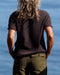 Person wearing a dark brown t-shirt and green pants standing by a body of water.