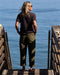 Man standing on a wooden dock by the water wearing a dark shirt and green pants.