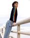 Person wearing a black sweater with rainbow stripes and blue jeans, standing on a wooden railing by the sea.