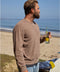 Man wearing brown sweater holding a bottle on a beach with ocean in the background