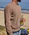 Man wearing brown sweater holding a bottle on a beach with ocean in the background