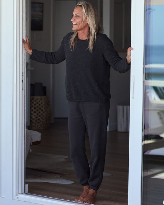 Woman standing by a door, wearing a black outfit, with a neutral background.