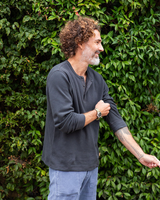 Man wearing a black long-sleeve shirt and light blue pants standing against a green leafy background