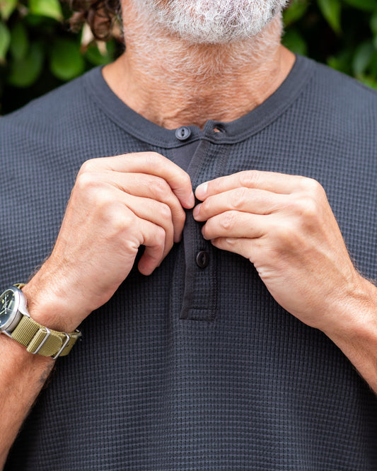 Man adjusting buttons on a black shirt with a blurred green background