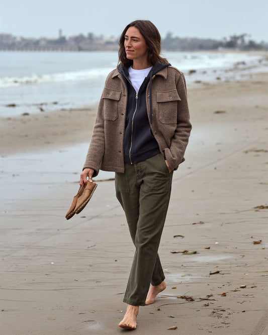 Person standing on a beach wearing a brown jacket and green pants, holding shoes.