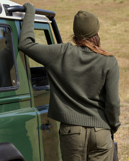 Person in green sweater and beanie standing next to a green vehicle in a field
