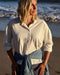 Woman standing on a beach wearing a white shirt and denim wrap, with ocean waves in the background.