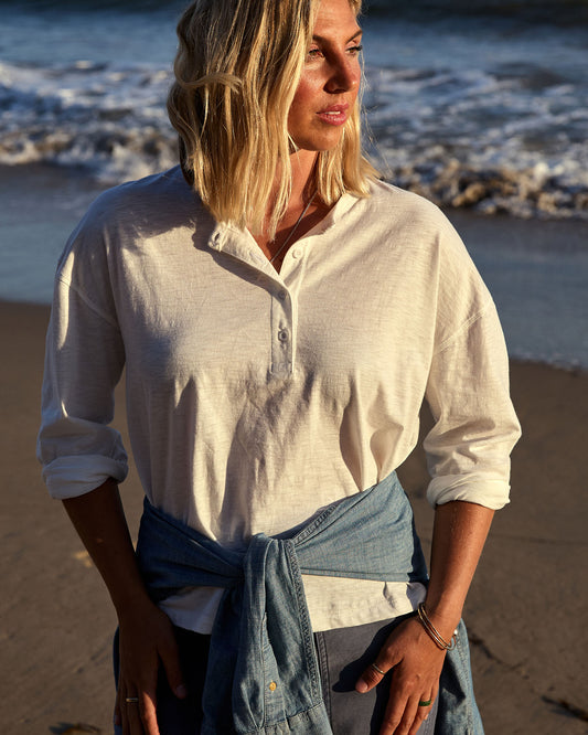 Woman standing on a beach wearing a white shirt and denim wrap, with ocean waves in the background.