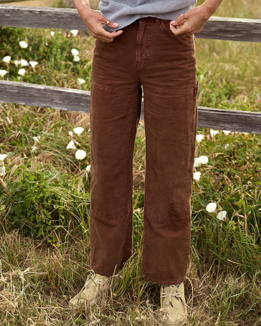 Person wearing brown corduroy pants standing in a field with white flowers and a wooden fence.
