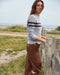 Woman wearing a gray sweater with colorful stripes and brown pants standing by a wooden post on a grassy area.