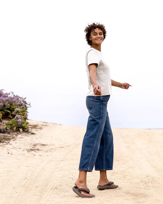 Person wearing a light grey t-shirt and blue jeans standing on a sandy beach with a plant in the background.
