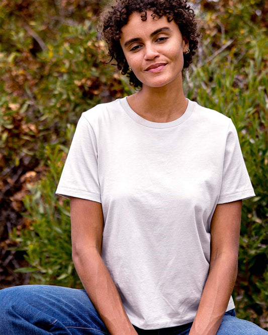 Person wearing a plain light grey t-shirt sitting outdoors with greenery in the background
