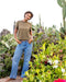 Person wearing a brown shirt and blue jeans standing in a garden with cacti and other plants.