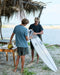 Two people on a beach with a surfboard, one holding a coconut.