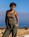 Person wearing a olive brown tank top and pants standing on a coastal landscape with ocean and sky in the background.