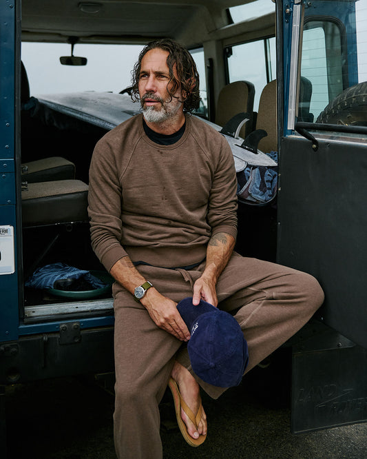Man sitting inside a vehicle holding a blue cap, wearing a brown sweater and sweatpants.