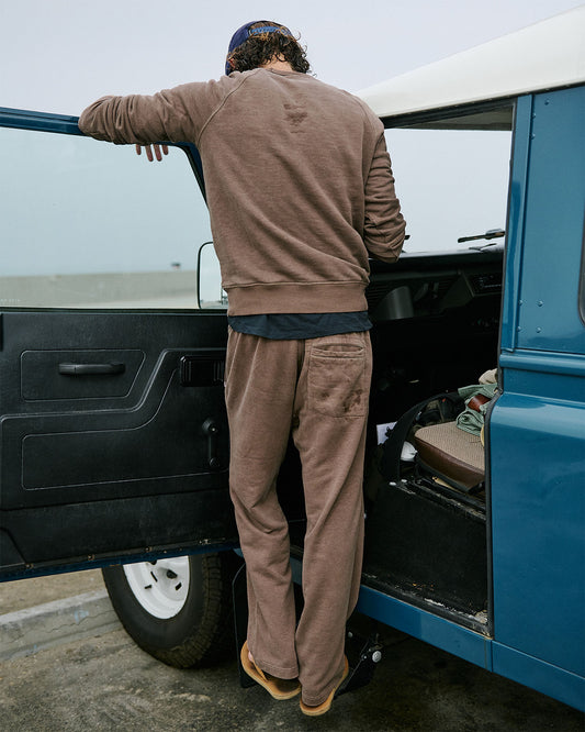 Man standing next to a blue vehicle with an open door, wearing a brown sweater and sweatpants.