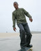 Man in green sweatshirt and black pants skateboarding on a concrete surface with a clear sky.