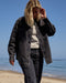 Person wearing a dark jacket and jeans walking on a beach with clear blue sky.