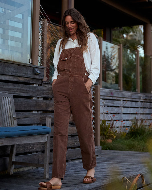 Woman wearing brown overalls standing on a wooden deck.
