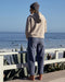 Person wearing a beige hoodie and blue pants standing on a wooden deck overlooking the ocean.