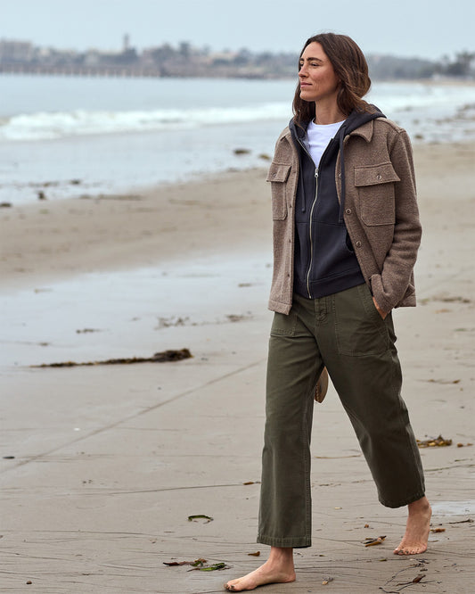 Person standing on a beach wearing a brown jacket and green pants.