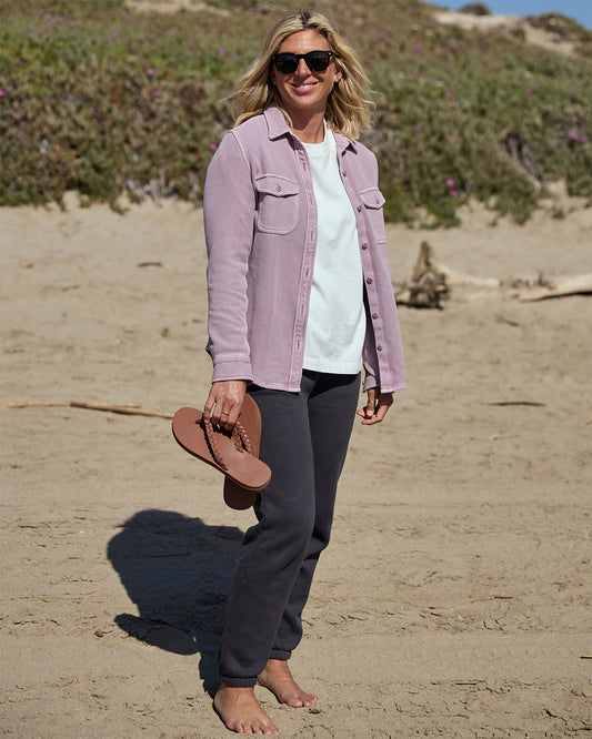 Woman standing on a sandy beach wearing a light pastel pink blanket shirt and dark pants, holding sandals.