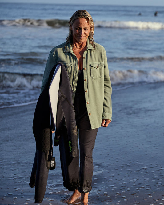 A woman wearing a green blanket shirt on the beach with ocean waves in the background