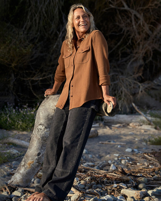 Person wearing a brown blanket shirt and dark pants standing on a rocky surface with dry vegetation in the background