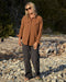 Person wearing a brown blanket shirt and dark pants standing on a rocky surface with dry vegetation in the background