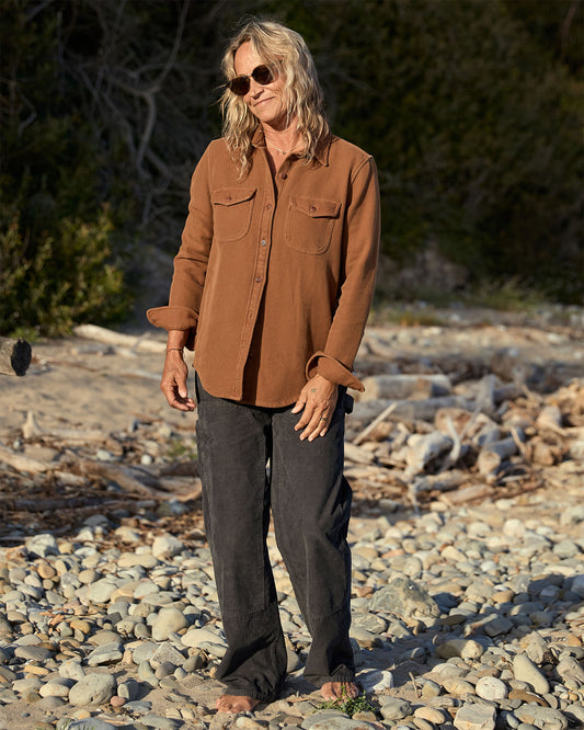 Person wearing a brown blanket shirt and dark pants standing on a rocky surface with dry vegetation in the background