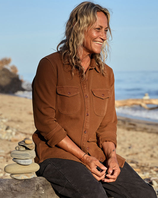 Person sitting on a beach with a stack of stones, wearing a brown blanket shirt.