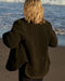 Person wearing a dark green wool jacket standing on a beach with ocean waves in the background