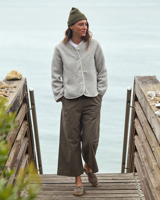 Woman standing on a wooden dock wearing a light gray wool jacket and brown pants with a green beanie.
