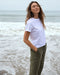 Woman wearing a white t-shirt and green pants standing on a beach.