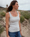 Woman wearing a white tank top and blue jeans standing on a sand path with a natural landscape in the background.