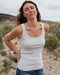 Woman wearing a white tank top standing on a beach with sand and grass in the background