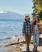 Two people walking along a lakeshore with mountains in the background